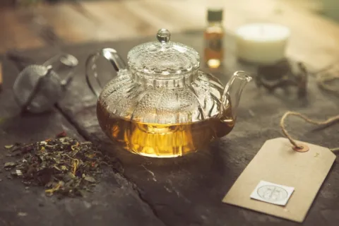 Glass teapot with herbal tea on a table, surrounded by loose leaves, a candle, and a tag.