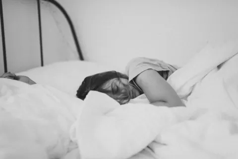 Person sleeping on a bed with white sheets in a minimalistic room.