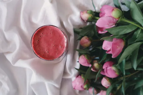 Pink smoothie in a glass next to pink peonies on white fabric.