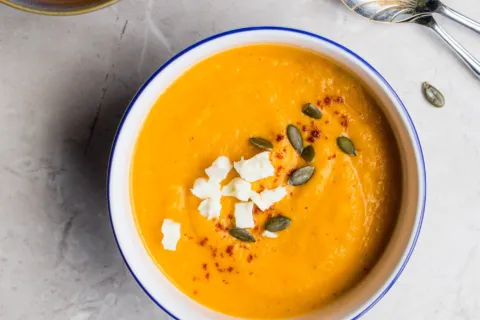 Two bowls of pumpkin soup with seeds and cheese on a marble surface.