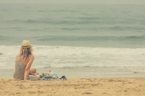 Woman in swimsuit and hat sitting on sandy beach, facing the sea.