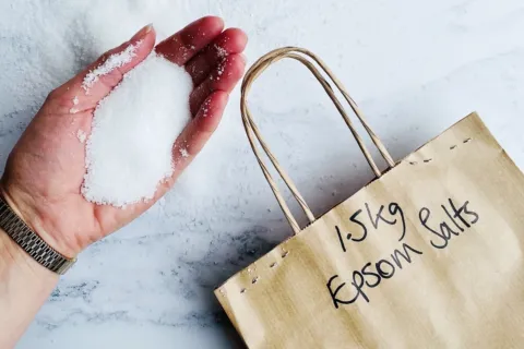 Hand holding Epsom salts next to a paper bag labelled "1.5kg Epsom Salts".