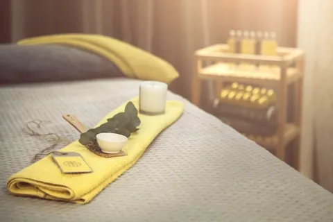 Yellow towel with candle, eucalyptus, and bowl on massage table. Shelves with bottles in background.