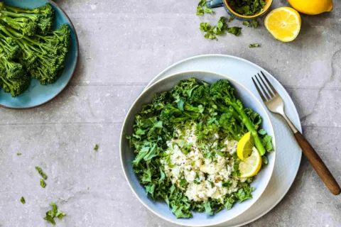 Bowl of rice with greens and lemon slices, fork on side, broccolini and lemon on table.