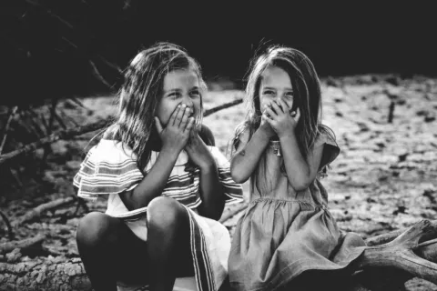 Two young girls sitting on a log, covering their mouths, outdoors.