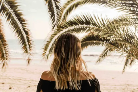 Woman with long hair facing the beach, surrounded by palm trees.