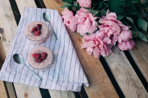 Two bowls of pink dessert with raspberries on a cloth, next to pink flowers on a wooden table.