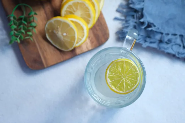 Glass of water with lemon slice, wooden board with lemon slices, and blue cloth.