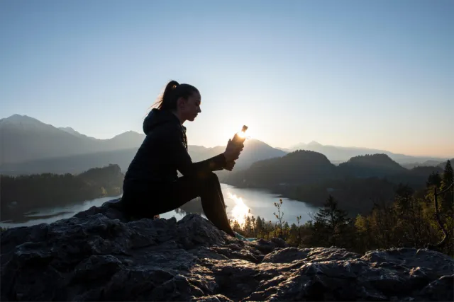 Person sitting on a rock at sunrise, holding a bottle, with mountains and lake in the background.