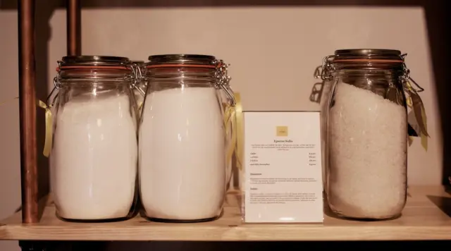 Three jars of Epsom salts on a wooden shelf with a product description card.