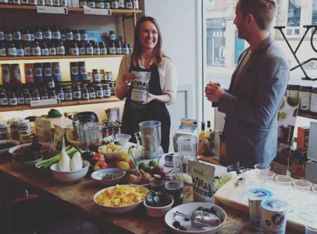 Two people at a table with fruits, jars, and a blender in a shop setting.