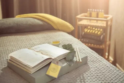 Open book, eucalyptus leaves, and candles on a spa bed with a yellow towel in a softly lit room.