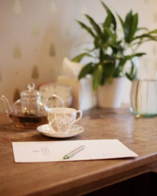 Teapot, teacup, and paper on wooden table with plant and water jug in background.