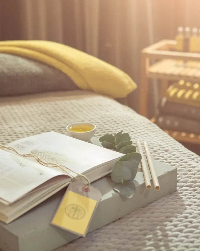 Open book, eucalyptus leaves, and candles on a spa bed with a yellow towel in a softly lit room.