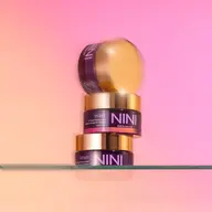 Three NINI Organics Violet beauty masks stacked on a glass shelf, pink background.