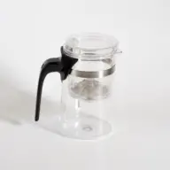 Clear glass teapot with black handle and metal band on white background.