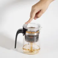 Hand pressing button on a glass coffee maker with black handle.