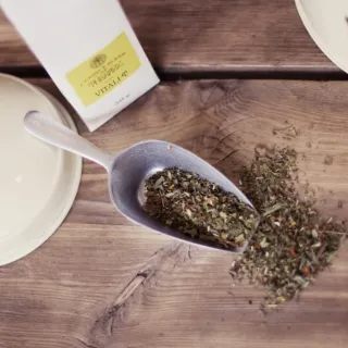 Herbal tea leaves in a metal scoop on a wooden table, with a Vitali-Tea packet nearby.