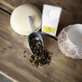 Loose tea leaves on a scoop, tea packet, and empty floral teacup on a wooden table.