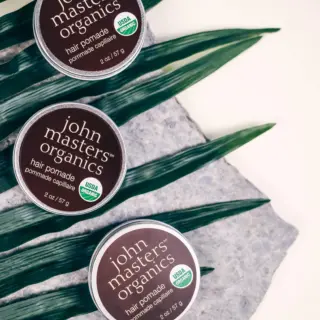 Three tins of John Masters Organics hair pomade on leaves and stone surface.