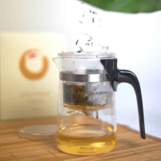 Glass teapot with infuser brewing tea, on a bamboo mat. Box with circular logo in background.