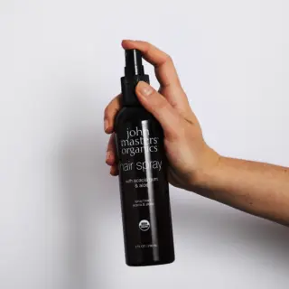 Hand holding John Masters Organics hair spray bottle against white background.