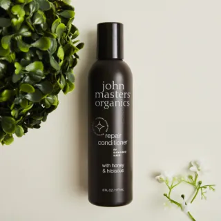 John Masters Organics repair conditioner bottle with honey and hibiscus, surrounded by green leaves.