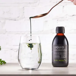 Liquid chlorophyll drops into water beside a Kiki Health bottle on a white surface.