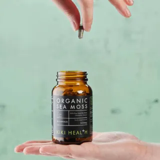 Hand holding a capsule above an open jar of Kiki Health Organic Sea Moss capsules.