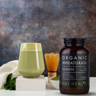 Organic wheatgrass powder jar, glass of green drink, whisk, and mint leaves on a white surface.