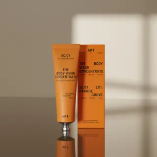 Orange tube and box of AKT SC.01 Body Wash Concentrate on reflective surface.