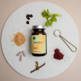 YourGut Glow supplement jar with herbs, seeds, capsules, and a spoon of salt on a round board.
