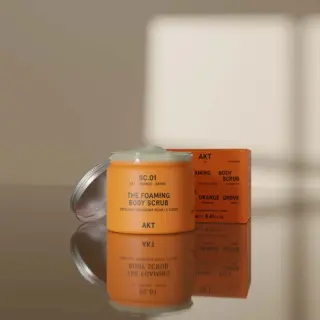 Orange AKT foaming body scrub jar and box on reflective surface.