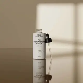 Open bottle of AKT Everyday Encore Body Oil with dropper on reflective surface.