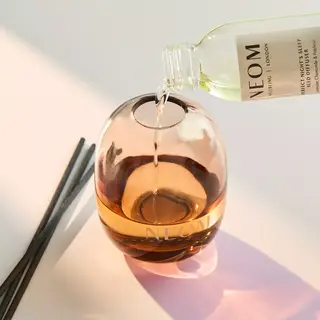 Diffuser oil being poured into a NEOM glass diffuser with reeds beside it.