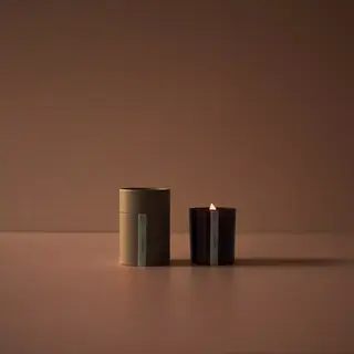 Lit candle in a dark glass holder next to a brown cylindrical box on a brown surface.