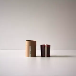 Brown candle box and lit red glass candle on white surface.