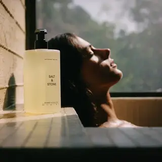 Bottle of Salt & Stone body wash on tiled ledge, person relaxing in bath by window.