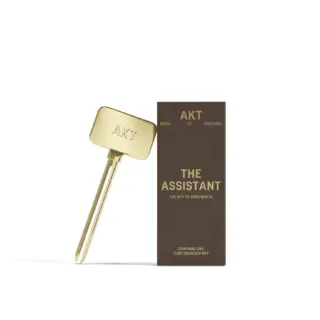 Gold tube squeezer key with "AKT" branding next to a brown box labelled "The Assistant".