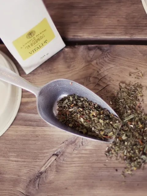 Herbal tea leaves in a metal scoop on a wooden table, with a Vitali-Tea packet nearby.