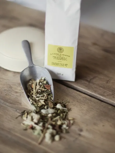 Herbal tea blend in a scoop on wooden table, with a white bag labelled "Immuni-Tea".