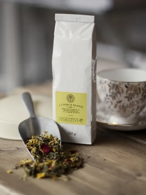 Herbal tea blend labelled "Digestivi-T" with a scoop and floral teacup on a wooden table.