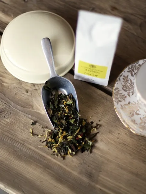 Loose tea leaves on a scoop, tea packet, and empty floral teacup on a wooden table.