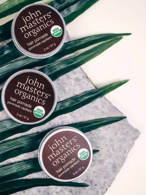 Three tins of John Masters Organics hair pomade on leaves and stone surface.