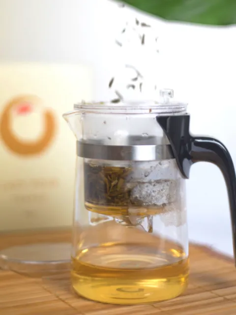 Glass teapot with infuser brewing tea, on a bamboo mat. Box with circular logo in background.