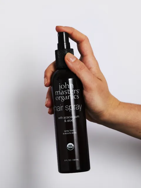 Hand holding John Masters Organics hair spray bottle against white background.