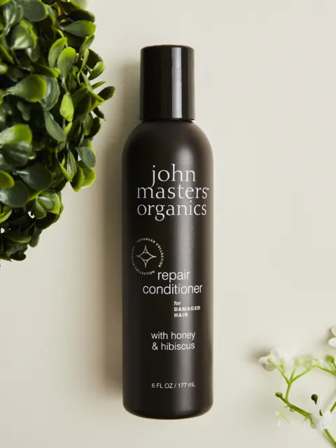 John Masters Organics repair conditioner bottle with honey and hibiscus, surrounded by green leaves.