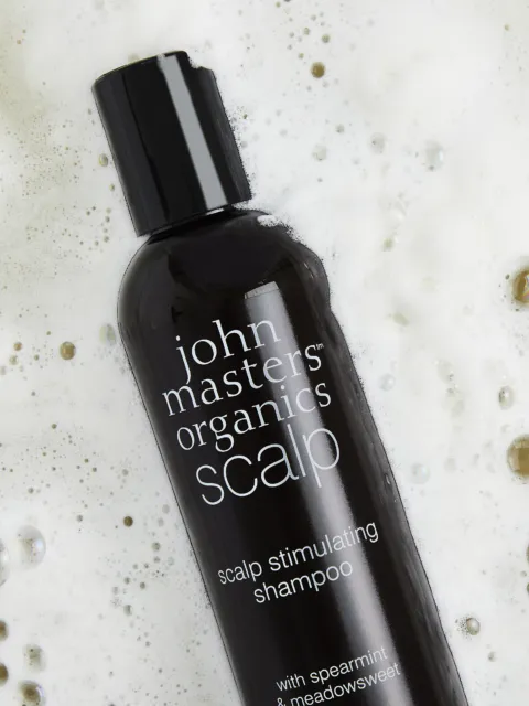 Black bottle of John Masters Organics scalp shampoo on foamy surface.