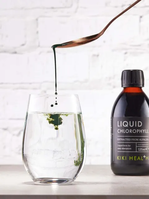 Liquid chlorophyll drops into water beside a Kiki Health bottle on a white surface.