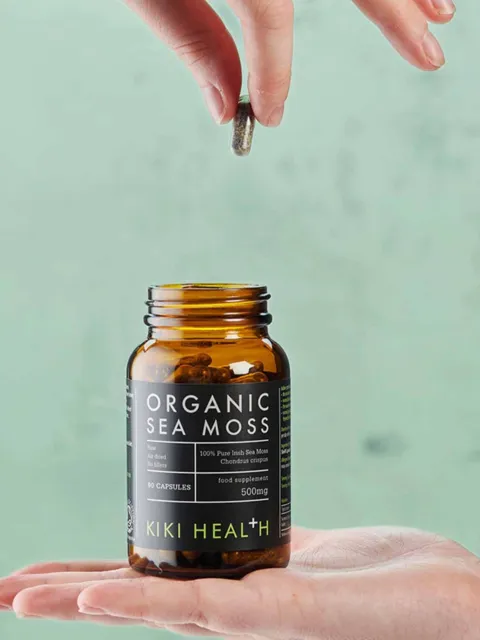 Hand holding a capsule above an open jar of Kiki Health Organic Sea Moss capsules.